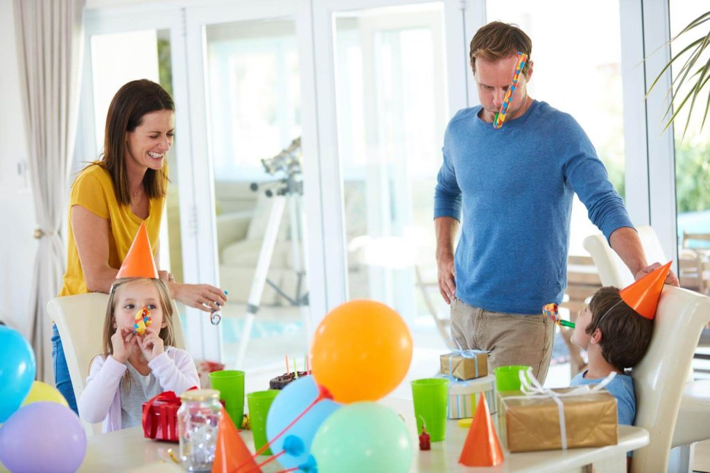 A family celebrates a child's birthday party at home with decorations, party hats, and a cake on the table.