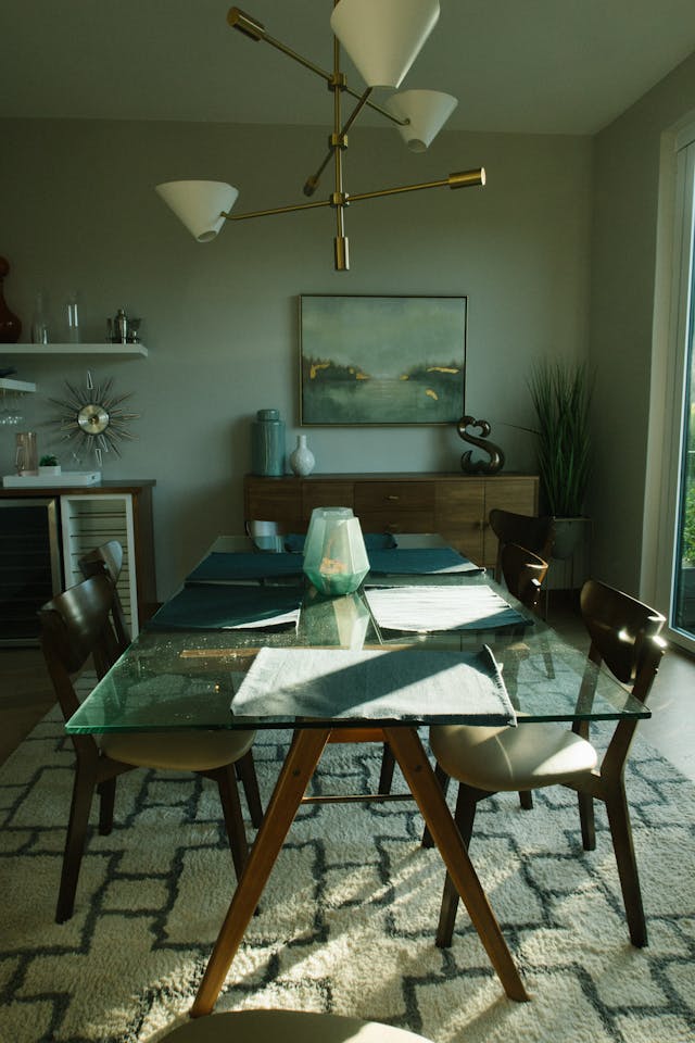 A modern dining room with a glass-top table, four wooden chairs, and a starburst chandelier. The walls are light grey, featuring a painting and decorative items on a sideboard and shelf.