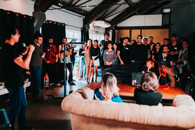 A group of people gathered in a room with a high ceiling, some standing and some sitting, while a person with a microphone speaks to the audience.