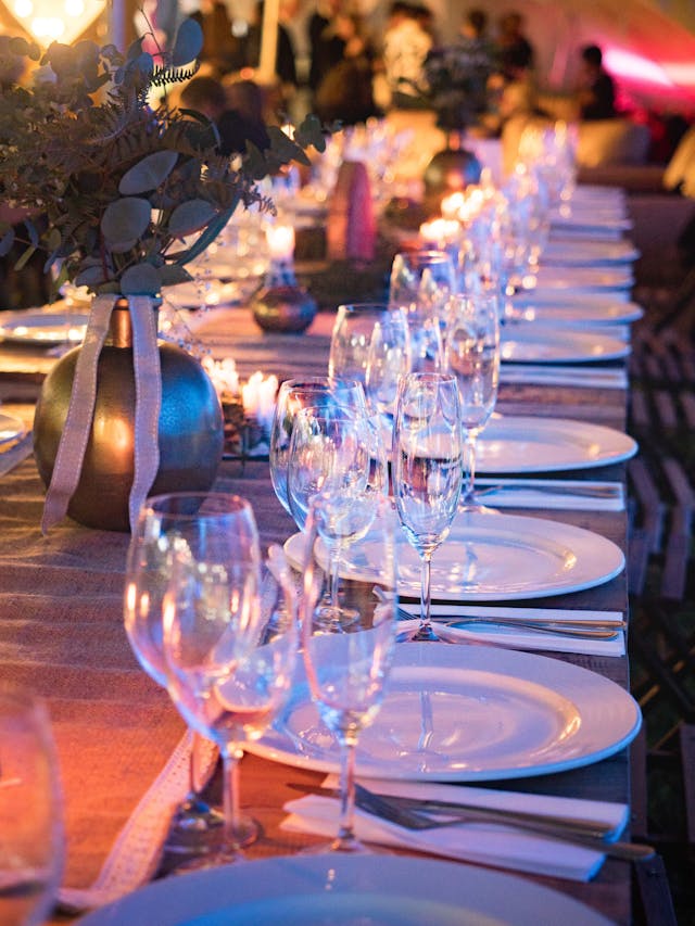 A long table set for a formal event with white plates, wine glasses, and a centerpiece under ambient lighting.