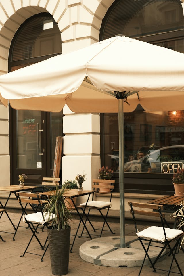 Outdoor café setting with empty tables and chairs beneath a large white umbrella. Potted plants are placed around the seating area, and there is a door with an "OPEN" sign in the background.