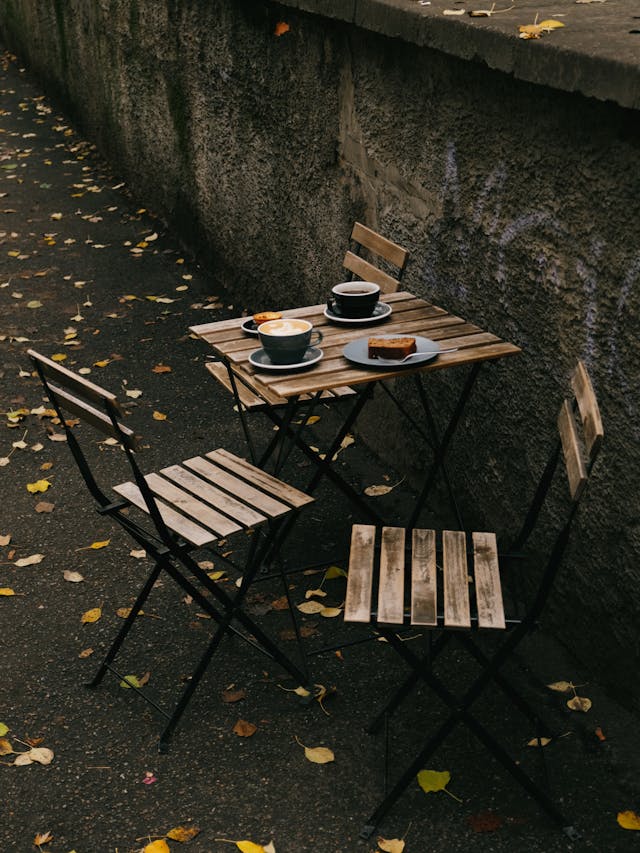 Two wooden chairs and a small wooden table with cups, saucers, and a plate on a leaf-strewn sidewalk next to a stone wall.