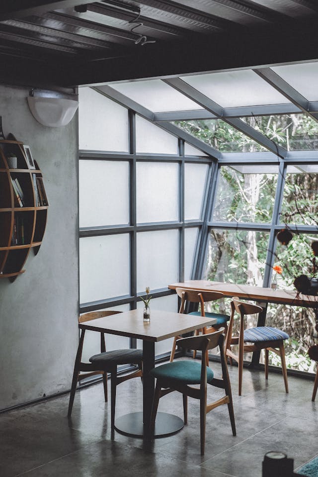 Modern cafe interior with wooden tables and chairs, large windows with a view of greenery outside, and minimalist decor.