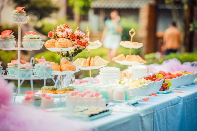 A table set with various desserts and finger foods, including cupcakes, sandwiches, fruits, and pastries. The setup is outdoors with a blurred background, featuring a few people and greenery.