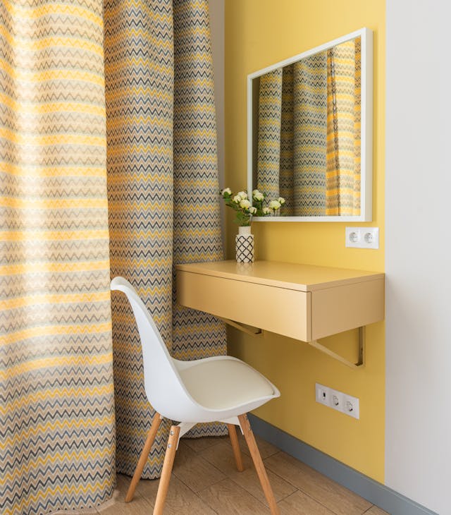 A small vanity with a wall-mounted yellow desk, white chair, and mirror. The corner is decorated with yellow patterned curtains and a small vase with flowers.