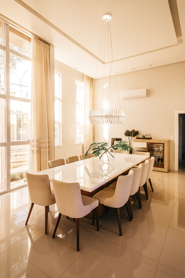 A modern dining room with a long rectangular table surrounded by white chairs. The room features large windows, beige curtains, a contemporary chandelier, and decorative plants.