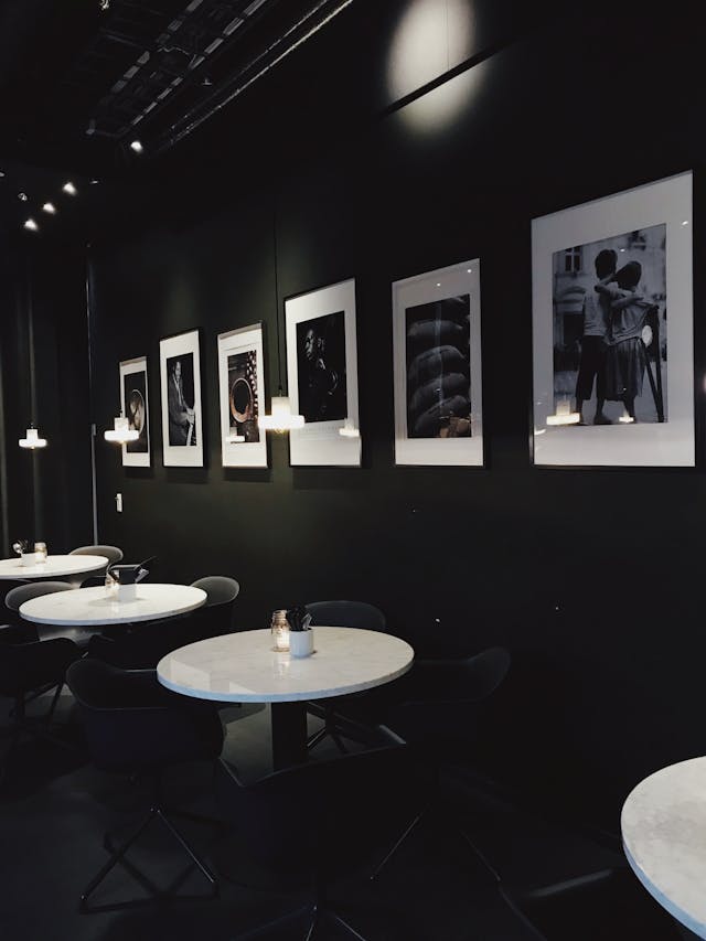 A dimly lit café interior with round white marble tables, black chairs, and black-and-white photographs on the dark walls.