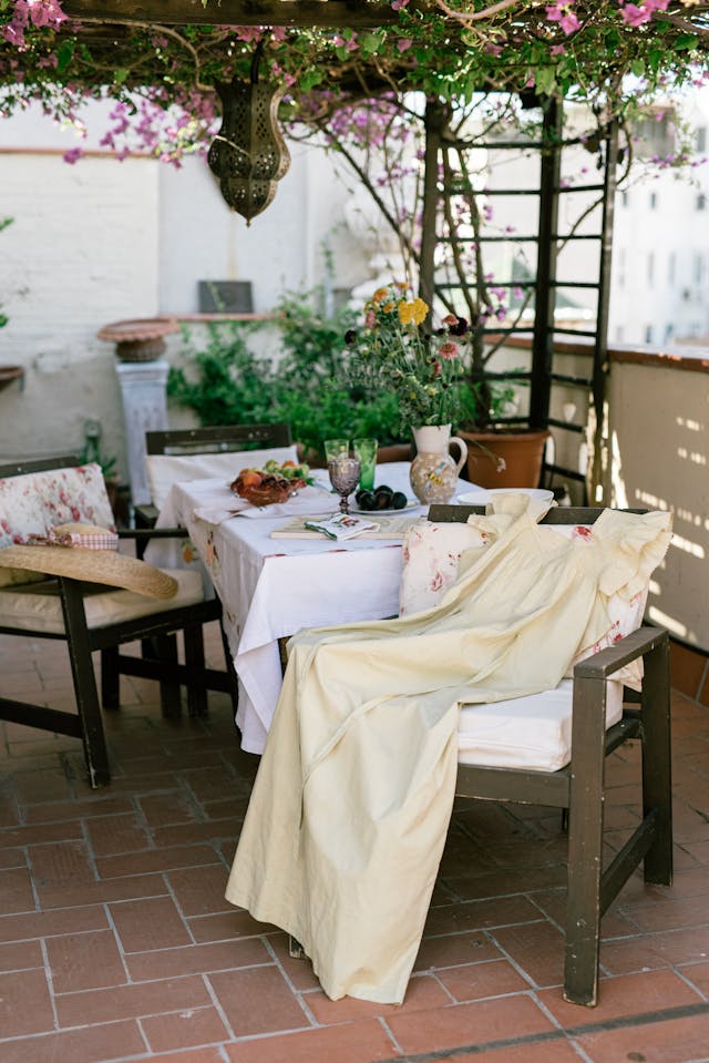 A patio dining area with a table covered by a white cloth and set for a meal, surrounded by chairs with cushions, underneath a pergola adorned with flowers and a lantern. A yellow dress is draped over one chair.