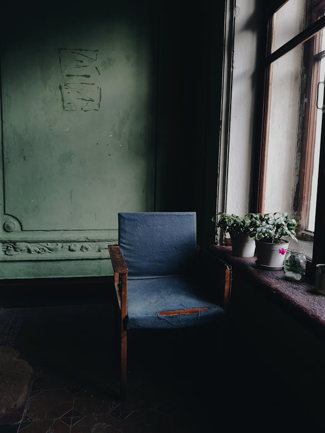 A worn blue chair is positioned next to a window with three potted plants on the windowsill. The room has dark green walls and appears dimly lit.