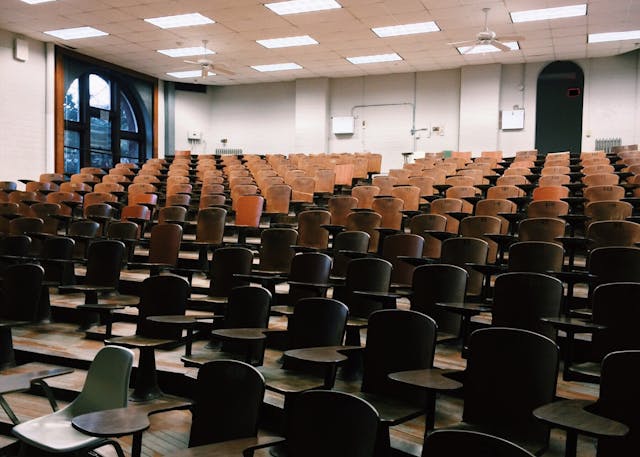 An empty lecture hall with rows of wooden desks and chairs. The room has a high ceiling with fluorescent lights and a large window on one side.