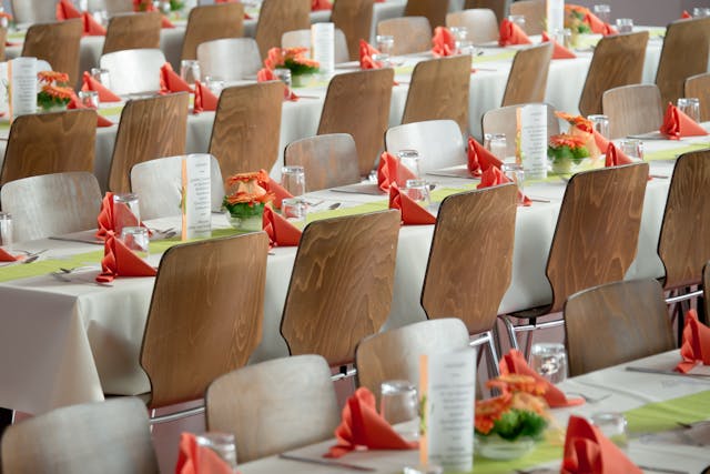 Rows of tables are set with green tablecloths, orange napkins, flower centerpieces, and menus, in a dining area with wooden chairs.