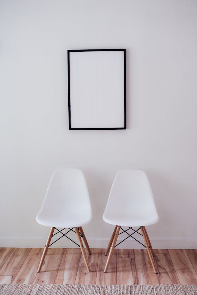 Two white modern chairs with wooden legs are placed side by side on a wooden floor against a blank white wall with an empty black frame above them.