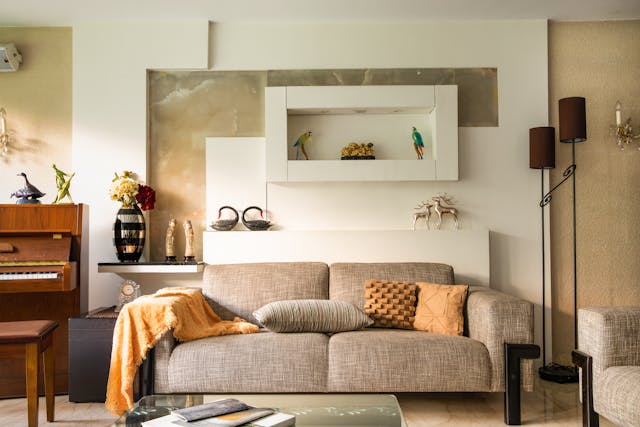 A modern living room with a gray sofa decorated with pillows and a throw. A piano is on the left, and wall art features bird figures. There are side tables, vases, and floor lamps on the right.