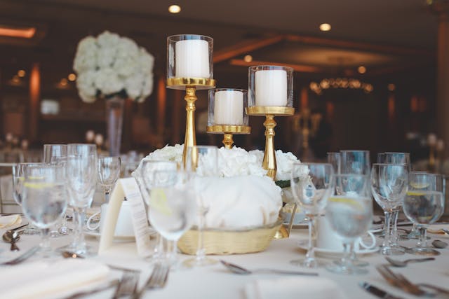 A table set for an event features glassware, cutlery, and white floral centerpieces with gold candle holders, each holding white candles.