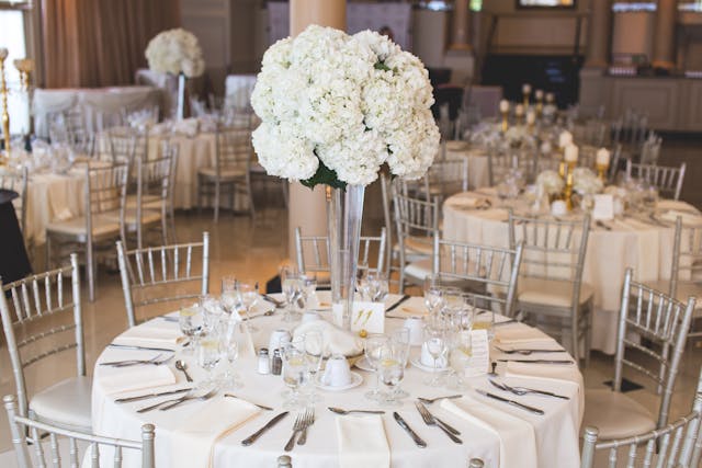 A dining table at a formal event is set with white linens, silverware, glassware, and a tall centerpiece of white flowers. Other similar tables are visible in the background.