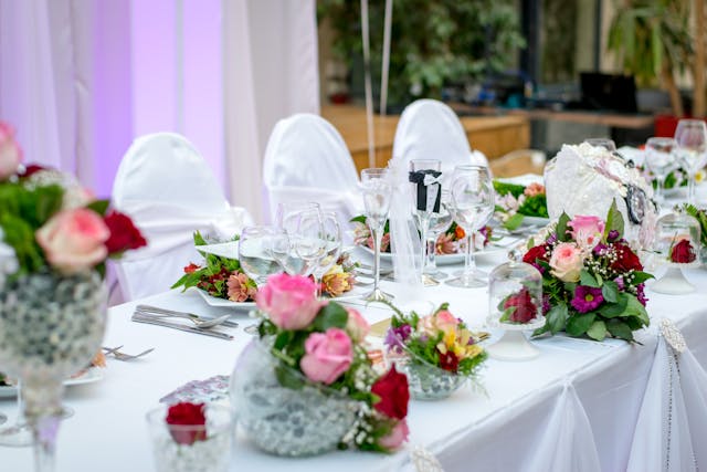 A wedding reception table is decorated with floral arrangements, white chair covers, and glassware.