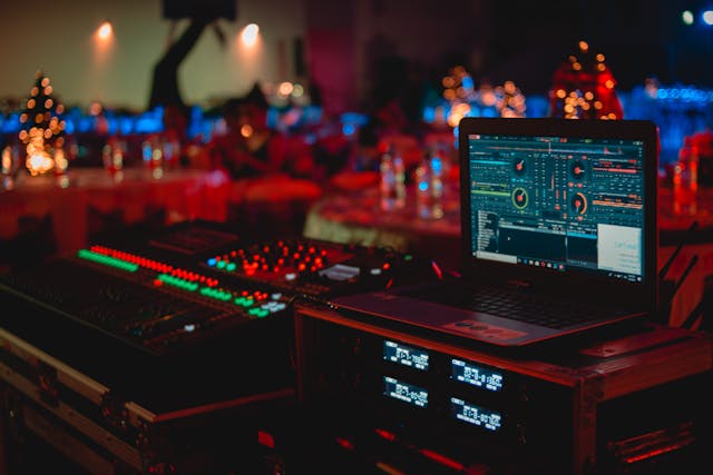 A laptop and DJ equipment with colorful LED lights are set up at a dimly lit event with tables and decorations in the background.