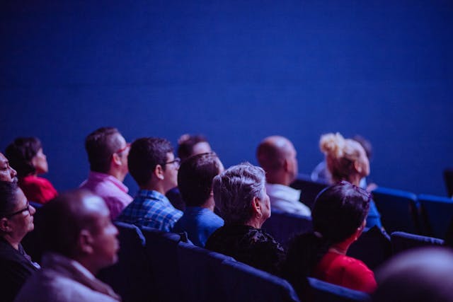 A group of people seated in rows, facing forward and attentively listening in an auditorium with a blue background.