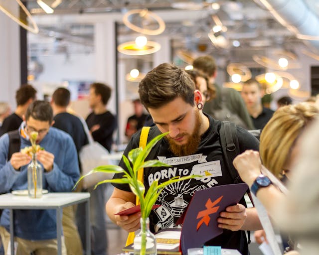 A group of people participate in an indoor event. A man in the foreground reads a pamphlet, while other attendees engage with displays and materials around the room.