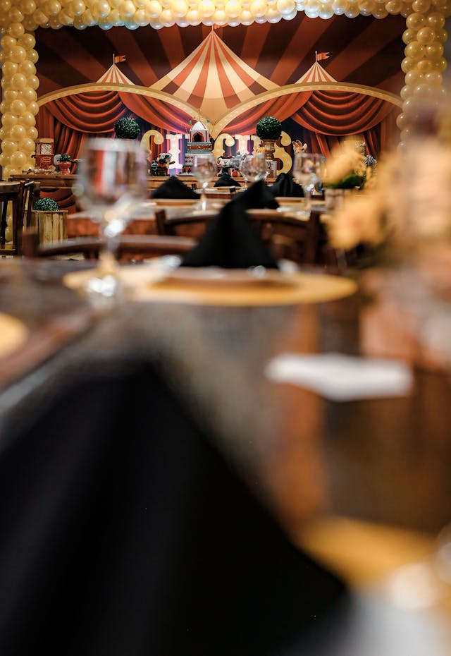 A dining table setup in focus with a blurred background showcasing a circus-themed stage with balloons and curtains.