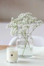 A Baby's Breathe in Glass containing white flowers next to a lit white candle on a white surface.