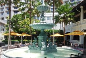 a large ornate fountain in an outdoor courtyard surrounded by palm trees and titan parasol yellow with seating areas and tall buildings visible in the background