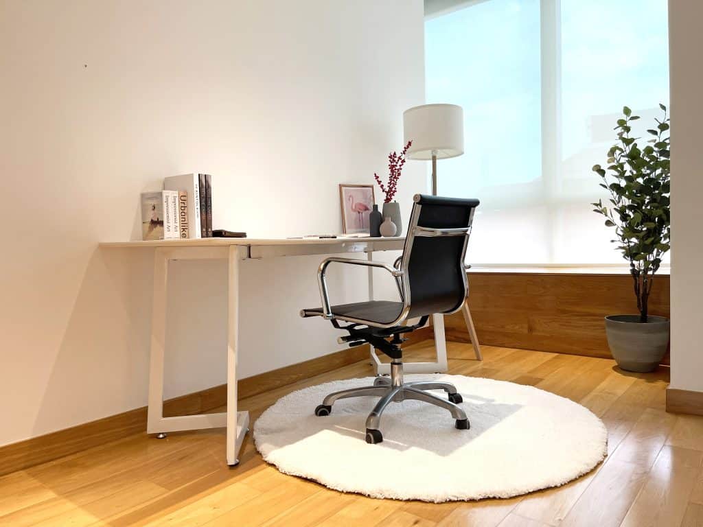 Minimalist home office with a white desk, black swivel chair, and lamp. A plant and books decorate the space, set on a light wood floor with a round rug.