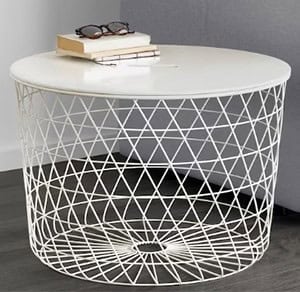 A round white table with a wireframe base sits on a floor next to a sofa. It holds a pair of glasses and two stacked books.