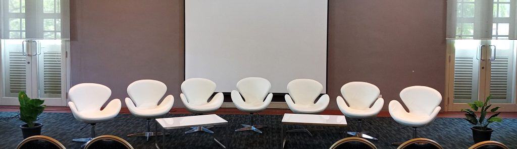 Seven white chairs arranged in front of a large blank screen, with small tables and plants on either side.