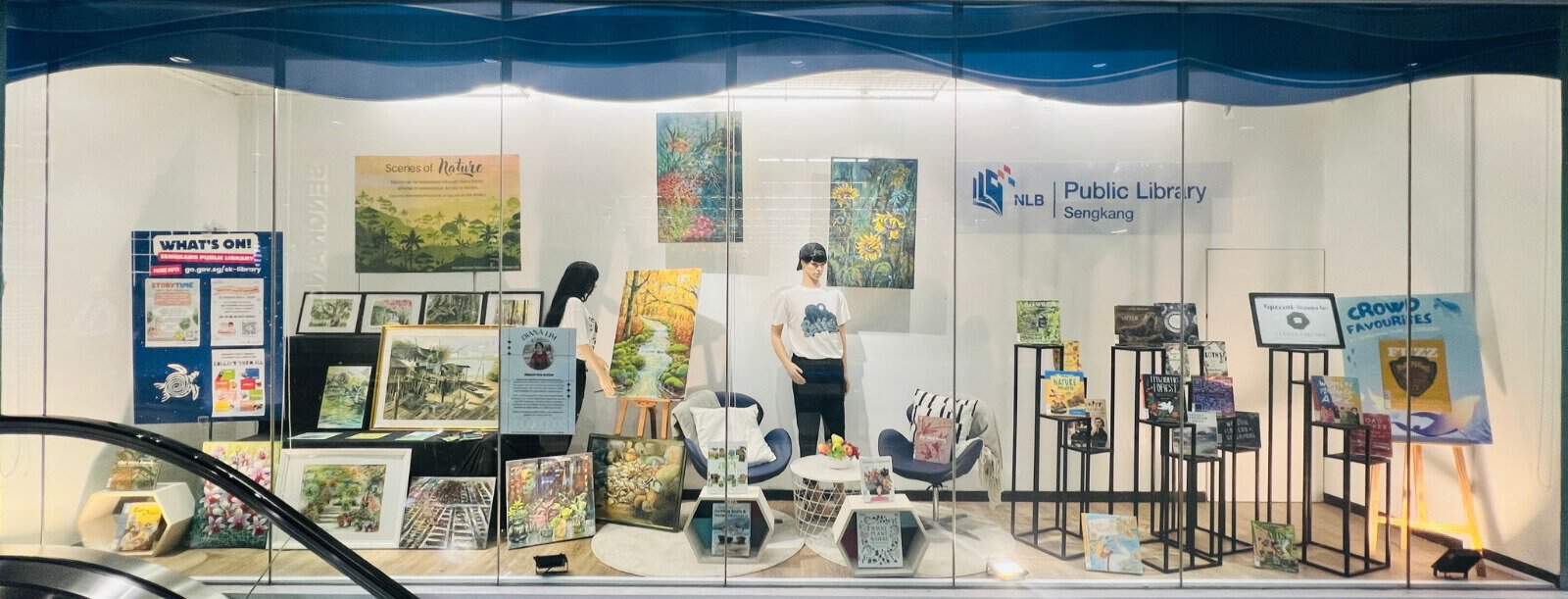 Library display window showcasing art and books, with paintings on easels, book racks, and a mannequin wearing a white shirt and sunglasses.