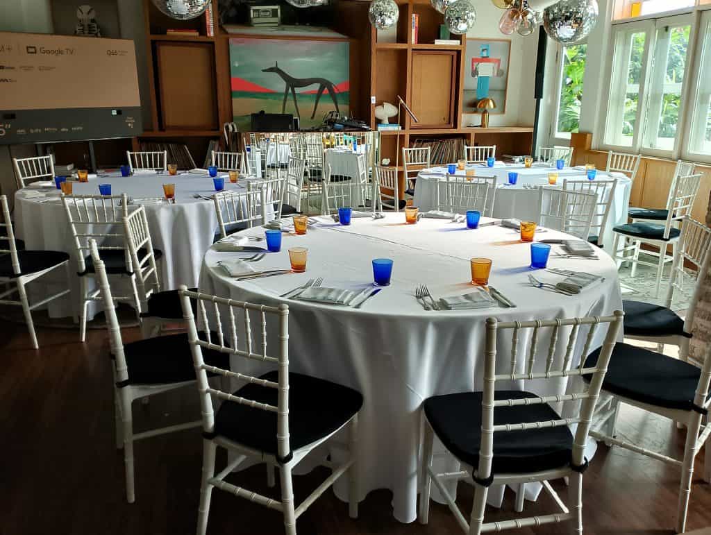 Round tables with white tablecloths and white chairs are set with orange and blue glasses in a bright room with large windows and a colorful painting.